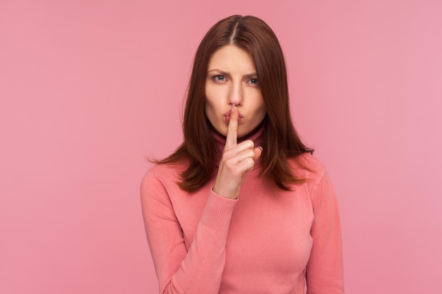 Mujer morena seria y preocupada que muestra un gesto shh sosteniendo el dedo cerca de los labios, guarda silencio, no hables. Disparo de estudio interior aislado sobre fondo rosa