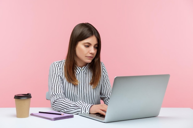 Mujer morena seria y segura de sí misma con camisa a rayas trabajando en una laptop sentada en un vaso de papel de oficina con una bebida y un bloc de notas acostado en el escritorio Foto de estudio interior aislada en fondo rosa