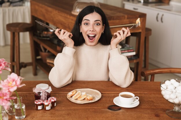 Foto mujer morena sincera desayunando y comiendo panqueques de queso con mermelada en la cocina de casa