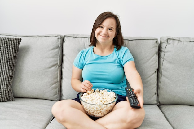 Mujer morena con síndrome de down sentada en el sofá viendo la televisión comiendo palomitas de maíz en la sala de estar