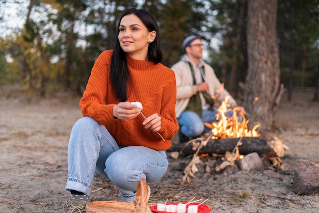 Mujer morena soñadora disfrutando de la recreación al aire libre mientras asaba malvaviscos sobre el fuego con su novio en el fondo. Foto de stock