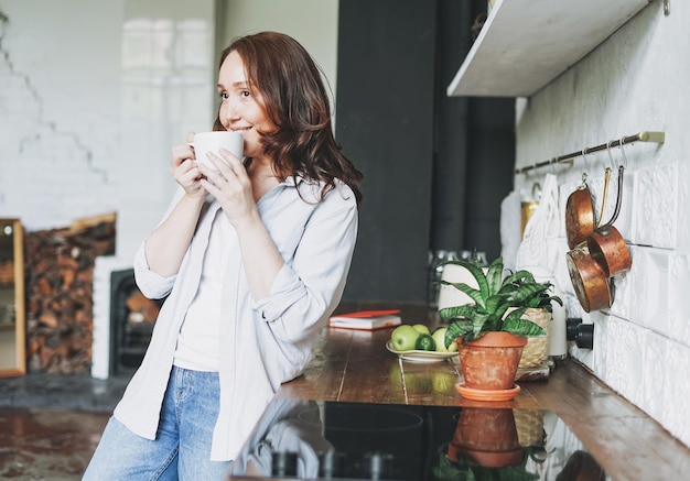 Mujer morena sonriente adulta en casual con una taza de té en la cocina en el hogar