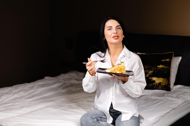 Mujer morena sonriente comiendo pastel en el dormitorio de su apartamento