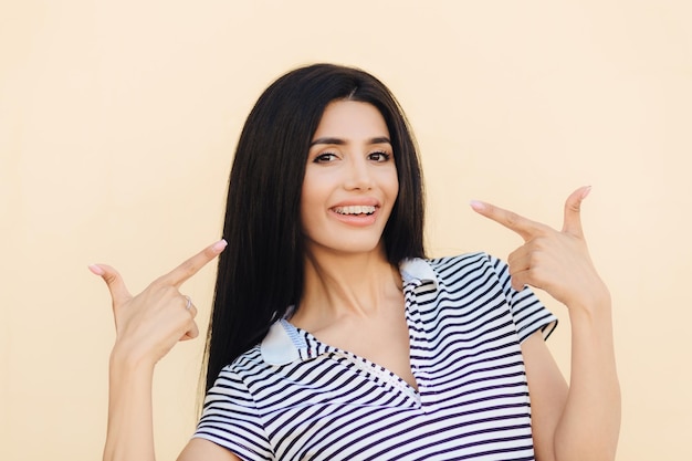 La mujer morena sonriente y complacida indica que en la boca tiene una expresión positiva indica que en la boca con una amplia sonrisa tiene el pelo largo y oscuro vestido con una camiseta a rayas aislada sobre un fondo rosa