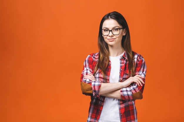 Mujer morena sonriente en lentes y presentación casual con los brazos cruzados y mirando a la cámara sobre fondo naranja.