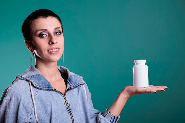 Foto mujer morena sonriente con el pelo corto sosteniendo una botella de medicamentos que presenta un producto sanitario a la cámara mientras está de pie sobre un fondo aislado en el estudio. complemento de revisión femenina alegre