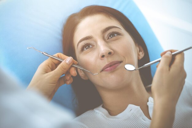 Una mujer morena sonriente siendo examinada por un dentista en una clínica dental. Las manos de un médico sosteniendo instrumentos dentales cerca de la boca del paciente. Dientes sanos y concepto de medicina.