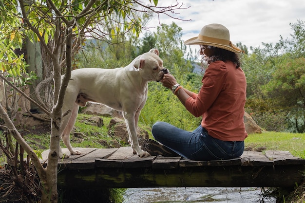 Mujer morena sonriente con sombrero alimentando a un perro sentado en un viejo puente de madera