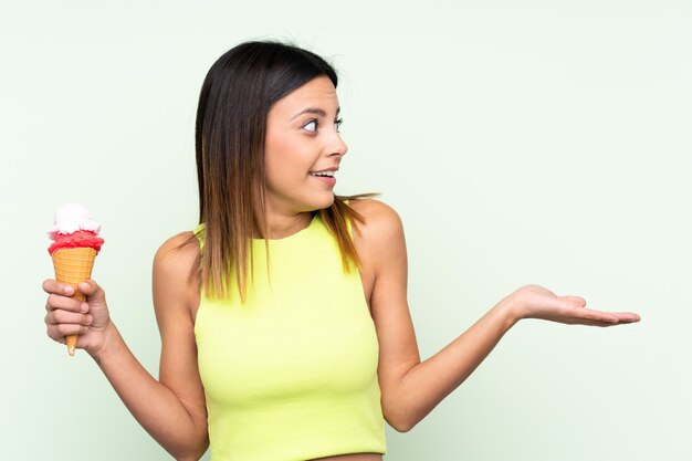 Mujer morena sosteniendo un helado de cucurucho sobre pared verde aislado con expresión facial sorpresa