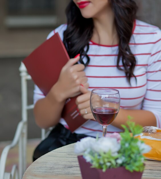 Mujer morena sosteniendo una revista y beber vino tinto en el café al aire libre.