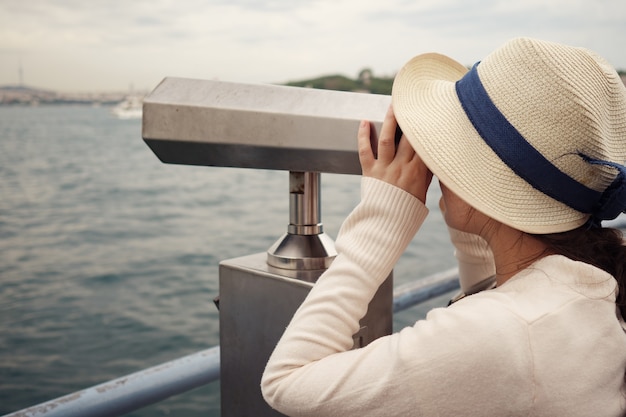 Mujer morena en suéter blanco y sombrero mira a través de binoculares explorando el paisaje urbano de cerca