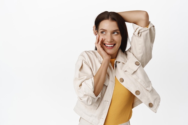 Foto mujer morena tierna y femenina, riendo coqueta, dientes blancos sonrientes, tocándose el pelo y las mejillas, sonrojándose tímidamente, mirando la pancarta de venta de la esquina superior izquierda, fondo blanco