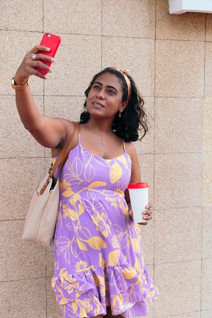 Mujer morena tomando un selfie con su teléfono inteligente y una taza de café en la calle