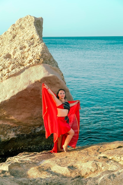 Una mujer morena con un traje para danzas orientales baila sobre una roca con el fondo del mar con un pañuelo.