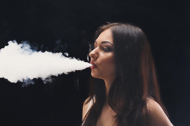 Mujer morena vaping cigarrillo electrónico con humo sobre fondo negro de cerca. Mujer joven fumando cigarrillo electrónico para dejar de fumar. Vapor y concepto alternativo de fumar sin nicotina, espacio de copia