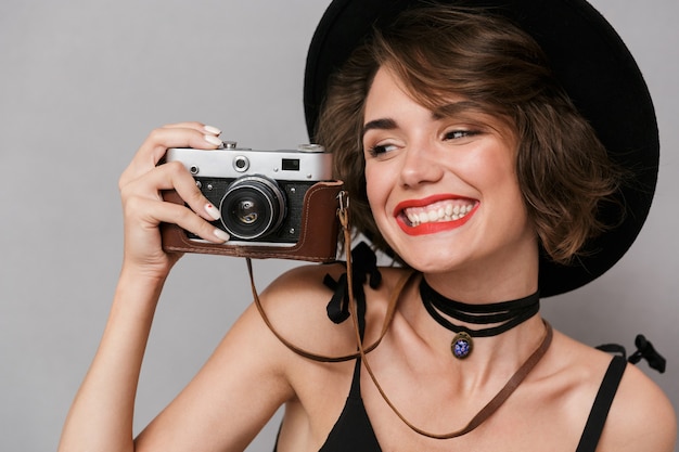 Mujer morena con vestido negro y sombrero fotografiando con cámara retro, aislado sobre pared gris