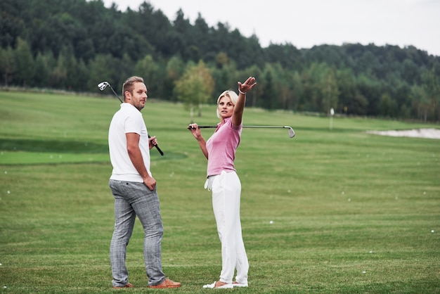 Mujer mostrando algo detrás. Par de jugadores de golf con palos en sus manos de pie sobre el césped.