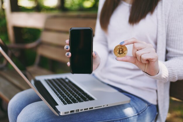 Mujer mostrando en la cámara del teléfono móvil con pantalla vacía en blanco para copiar el espacio. Mujer sentada en un banco sosteniendo bitcoin, moderno ordenador portátil al aire libre. Oficina móvil, concepto de moneda virtual en línea.