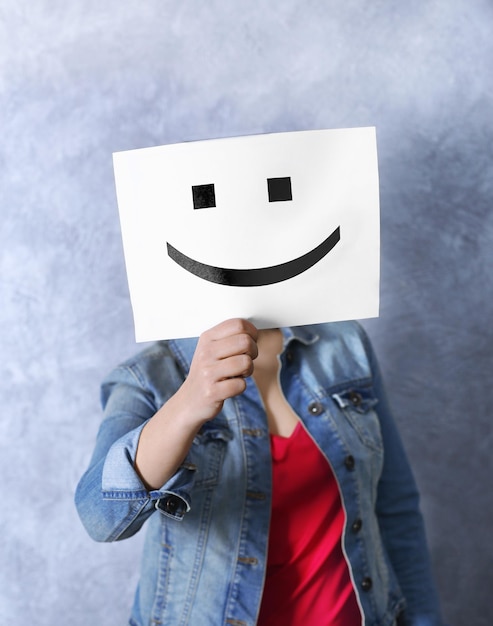 Mujer mostrando un emoticon feliz frente a la cara contra un fondo de pared de textura gris