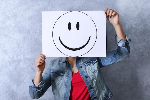 Mujer mostrando un emoticono feliz frente a la cara contra un fondo de pared con textura gris