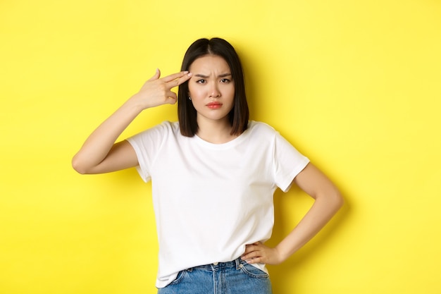 Mujer mostrando gesto de matarme, por favor, disparándose a sí misma en la cabeza con una pistola de dedo por aburrimiento, de pie molesta por el amarillo.