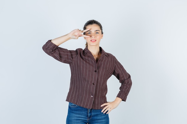 mujer mostrando el signo V cerca del ojo en camisa, jeans y luciendo orgullosa. vista frontal.