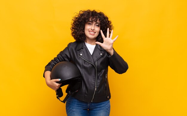 Mujer motociclista sonriendo y mirando amistosamente, mostrando el número cinco o quinto con la mano hacia adelante, contando hacia atrás