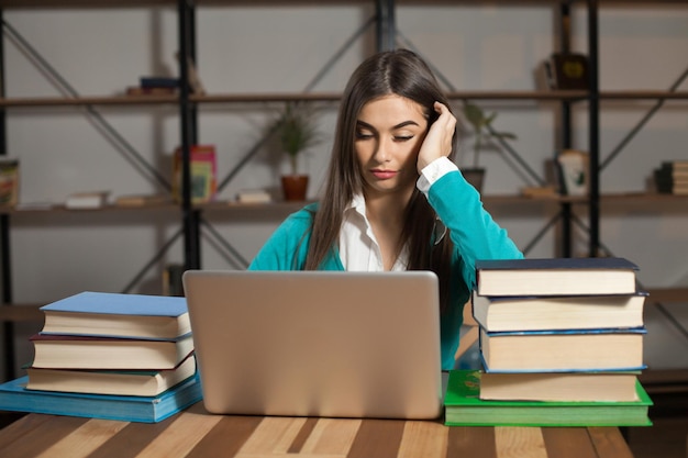 Una mujer con muchos libros y una computadora portátil está sentada en la mesa de madera