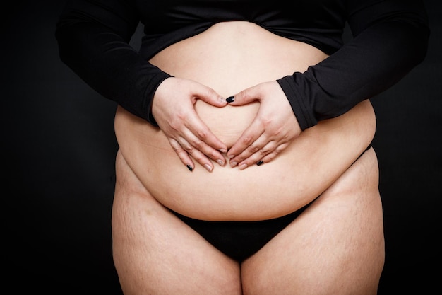 Una mujer muestra un corazón con las manos en una barriga gorda sobre un fondo negro concepto de positividad corporal de persona obesa Foto de alta calidad
