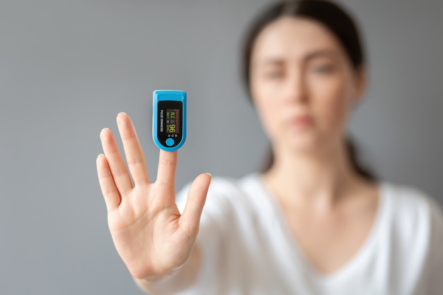 Una mujer muestra su mano con un pulsioxímetro en su dedo índice. Retrato borroso. Fondo azul. El concepto de medir el oxígeno en la sangre.