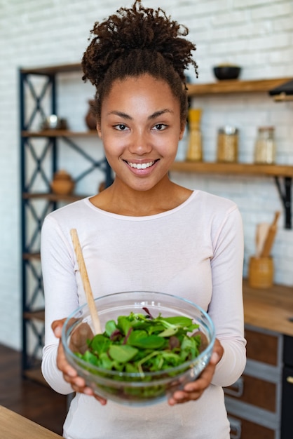 Mujer mulata bonita en casa mostrando comida saludable, sostiene en sus manos una ensalada