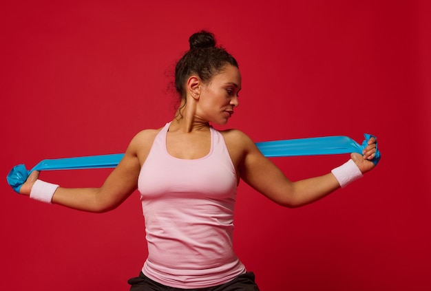 Mujer musculosa afroamericana haciendo ejercicios de estiramiento en los brazos usando banda elástica de fitness contra fondo rojo con espacio publicitario de copia