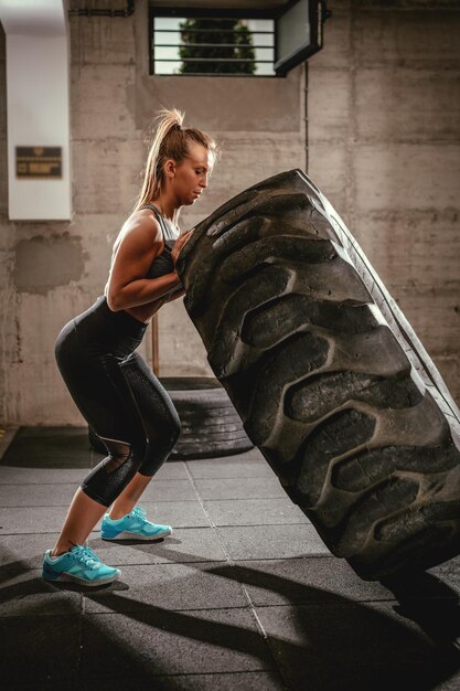 Mujer musculosa joven volteando un neumático en entrenamiento cruzado en el gimnasio.