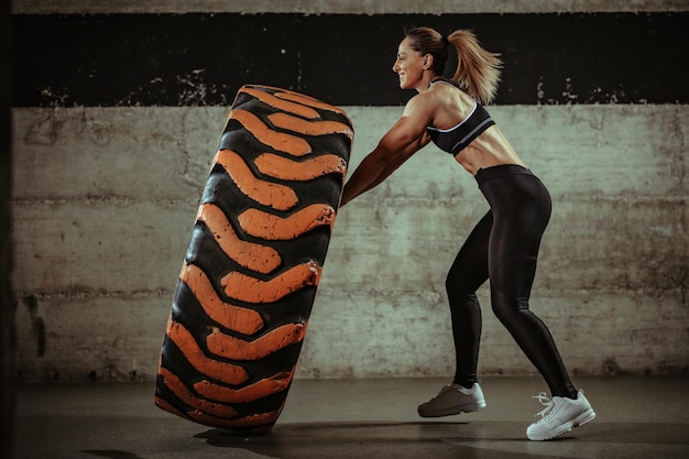 Mujer musculosa joven volteando un neumático de rueda en el entrenamiento en el gimnasio.