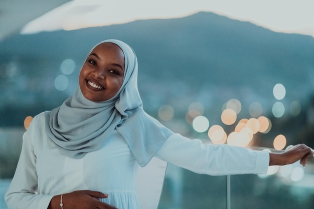 Mujer musulmana africana en la noche en un balcón sonriendo a la cámara con luces bokeh de la ciudad en la b