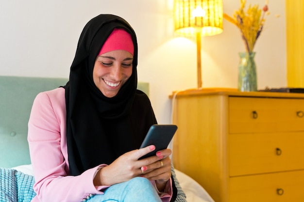 Mujer musulmana alegre que lleva un hijab con smartphone en interiores. Vista lateral horizontal de la mujer árabe con tecnología en la cama en casa. Tecnología y estilo de vida de las mujeres musulmanas.
