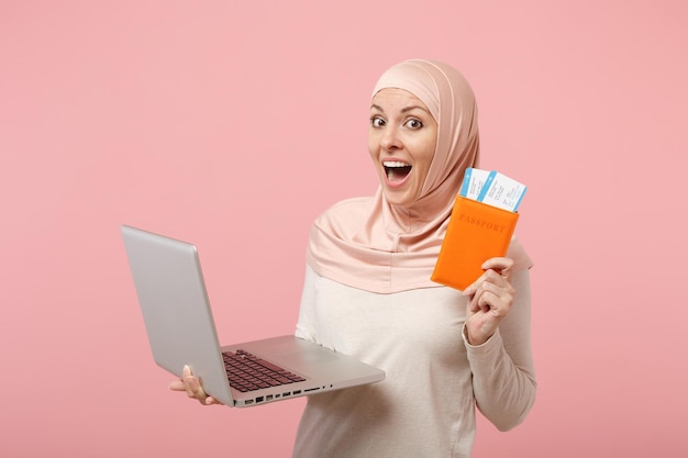 Mujer musulmana árabe emocionada con ropa ligera hijab posando aislada en un fondo rosa. Concepto de estilo de vida del Islam religioso de la gente. Simulacros de espacio de copia. Sosteniendo computadora portátil, pasaporte y boleto.