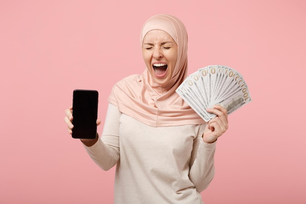 Mujer musulmana árabe feliz con ropa ligera hiyab posando aislada en un fondo rosa. Concepto de estilo de vida religioso de la gente. Simulacros de espacio de copia. Sostenga el teléfono celular con la pantalla vacía en blanco, fanático del dinero en efectivo.