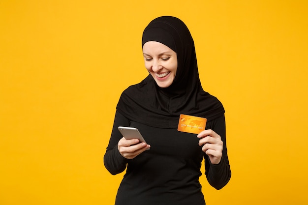 Mujer musulmana árabe joven en ropa negra hijab mantenga en la mano el teléfono móvil, tarjeta de crédito aislada en el retrato de la pared amarilla. Concepto de estilo de vida religioso de la gente.