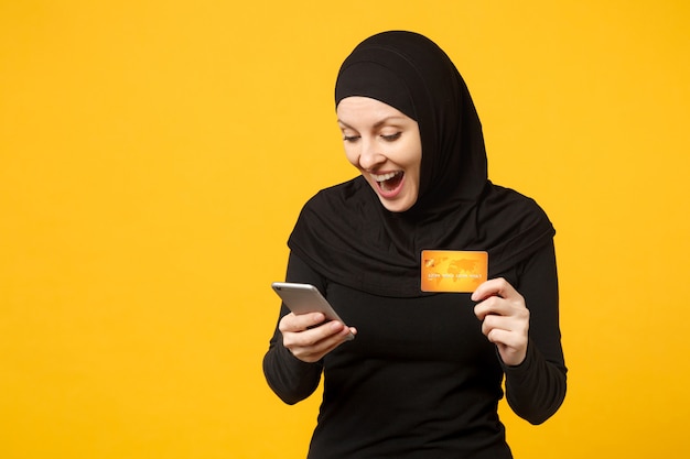 Mujer musulmana árabe joven en ropa negra hijab mantenga en la mano el teléfono móvil, tarjeta de crédito aislada en el retrato de la pared amarilla. Concepto de estilo de vida religioso de la gente.