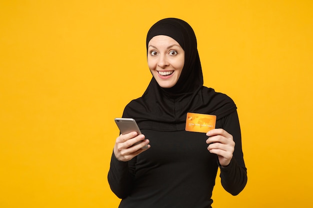 Mujer musulmana árabe joven en ropa negra hijab mantenga en la mano el teléfono móvil, tarjeta de crédito aislada en el retrato de la pared amarilla. Concepto de estilo de vida religioso de la gente.