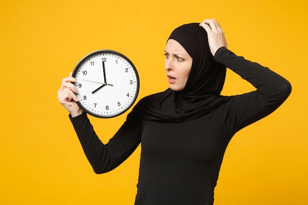 Mujer musulmana árabe joven en ropa negra hijab mantenga en manos reloj redondo aislado en retrato de pared amarilla. Estilo de vida religioso de la gente, concepto de gestión del tiempo.