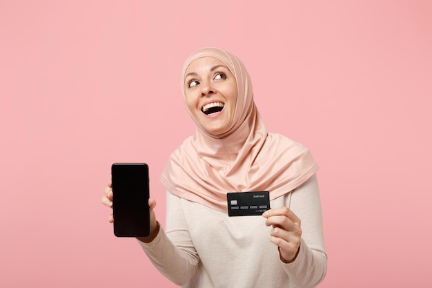 Mujer musulmana árabe pensativa con ropa ligera hiyab posando aislada en un fondo rosa. Concepto de estilo de vida religioso de la gente. Simulacros de espacio de copia. Sostenga el teléfono celular con una tarjeta bancaria de crédito en pantalla vacía en blanco.