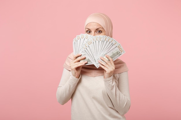 Mujer musulmana árabe con ropa ligera hiyab posando aislada de fondo rosa. Concepto de estilo de vida del Islam religioso de la gente. Simulacros de espacio de copia. Cara cubierta con abanico de dinero en efectivo en billetes de dólar.