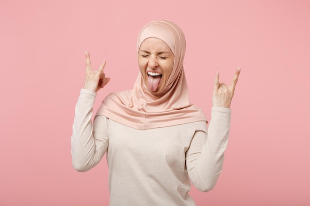 Mujer musulmana árabe con ropa ligera hiyab posando aislada de fondo rosa. Concepto de estilo de vida religioso de la gente. Simulacros de espacio de copia. Mostrando la lengua que representa el gesto de cuernos de letrero de rock pesado.