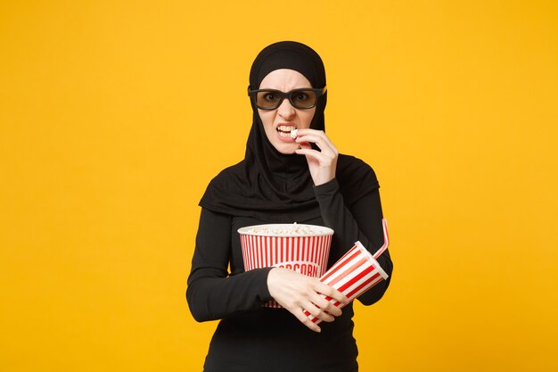 Mujer musulmana árabe en ropa negra hijab gafas imax 3d ver película con palomitas de maíz, taza de refresco aislado en retrato de pared amarilla. Concepto de estilo de vida de personas. .