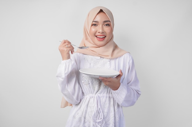 Foto mujer musulmana asiática joven hambrienta con hijab sosteniendo una cuchara y comiendo de un plato vacío con espacio de copia sobre el concepto de ramadán