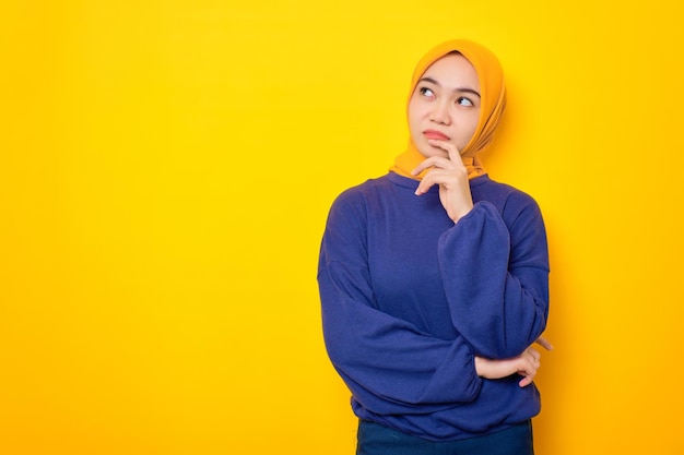 Foto mujer musulmana asiática joven y pensativa vestida con un suéter casual mirando el espacio de la copia y tocando la barbilla pensando en una oferta interesante aislada sobre fondo amarillo