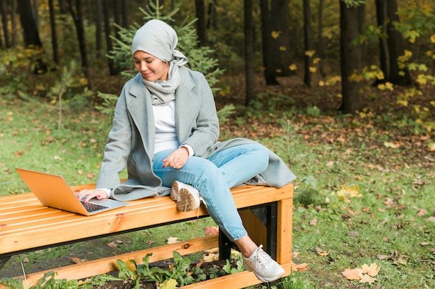 Mujer musulmana asiática joven que usa la computadora portátil en las compras en línea y sociales de la educación en línea del parque del otoño