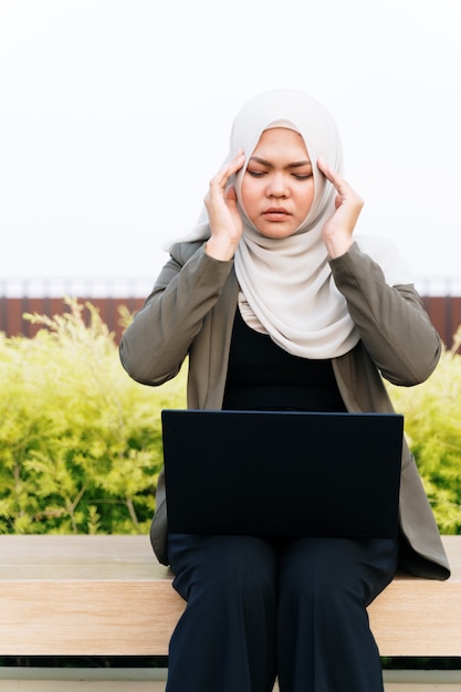 Mujer musulmana asiática joven subrayada en traje verde y trabajando en una computadora en el parque